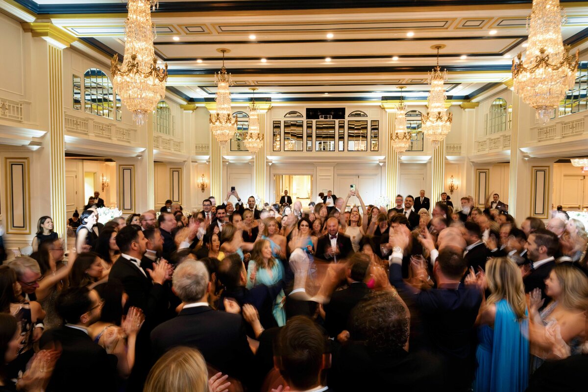 A large group of people in formal attire dance energetically in a grand, elegant ballroom with chandeliers and ornate decor. The room is filled with motion and celebration.