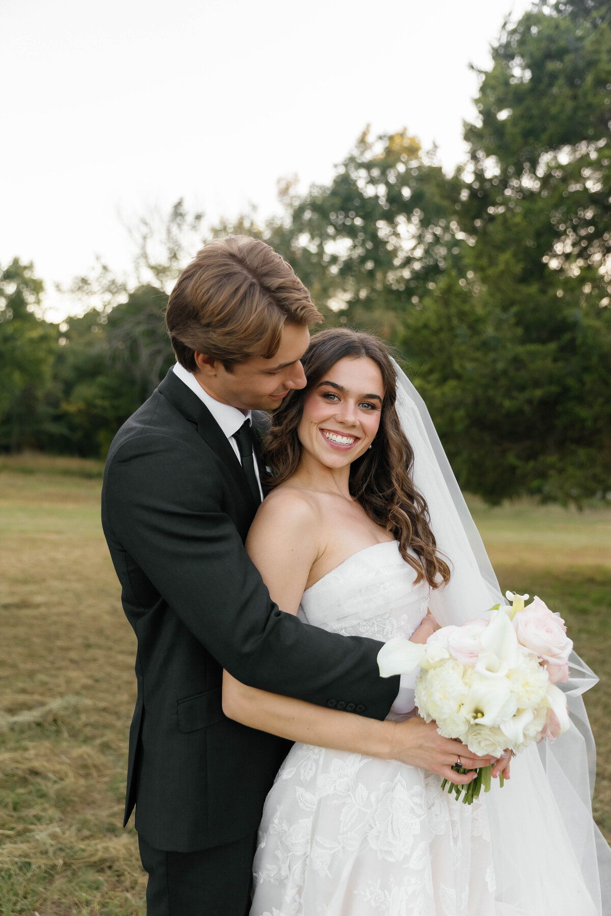 Bride and groom embracing in a sunlit field with the bride holding a full blush and ivory bouquet featuring garden roses and calla lilies designed by a fine-art Arkansas wedding florist.