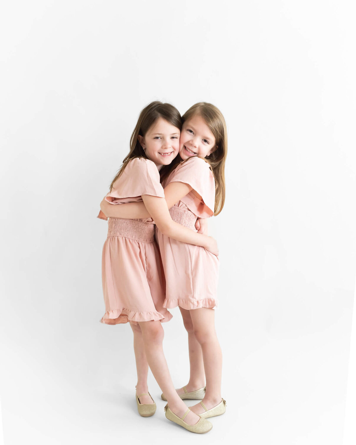 two young sisters in pink dresses giving eachother a big hug in studio session with white backdrop