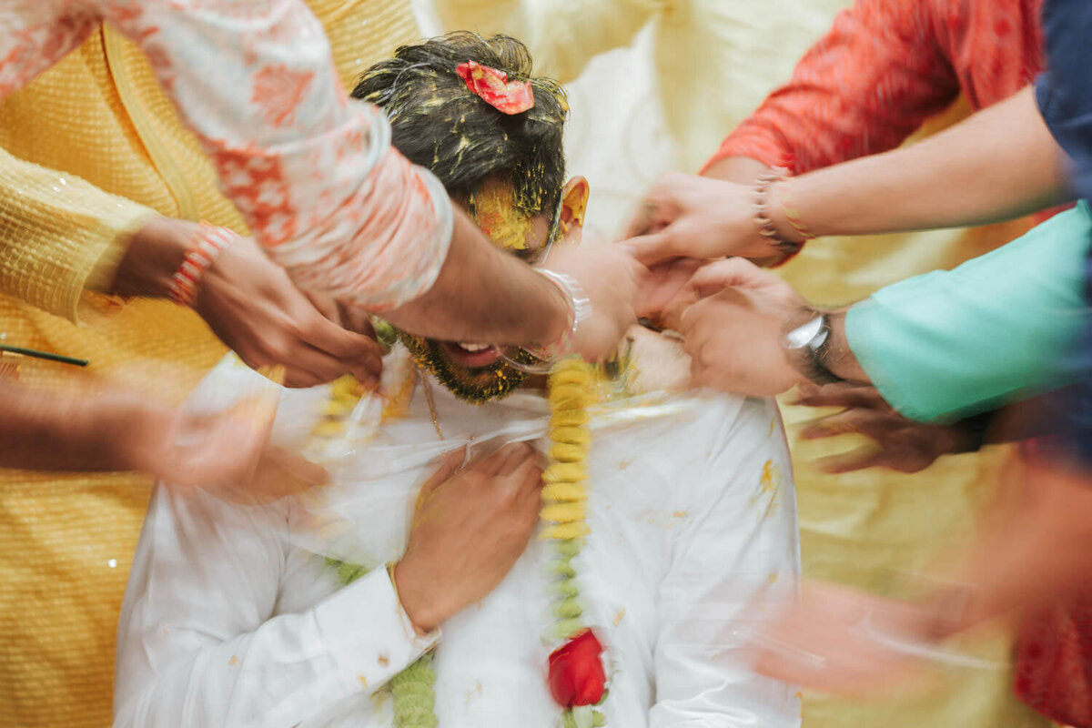 Friends and family surround a groom during a traditional haldi ceremony, applying turmeric paste with joyful enthusiasm.