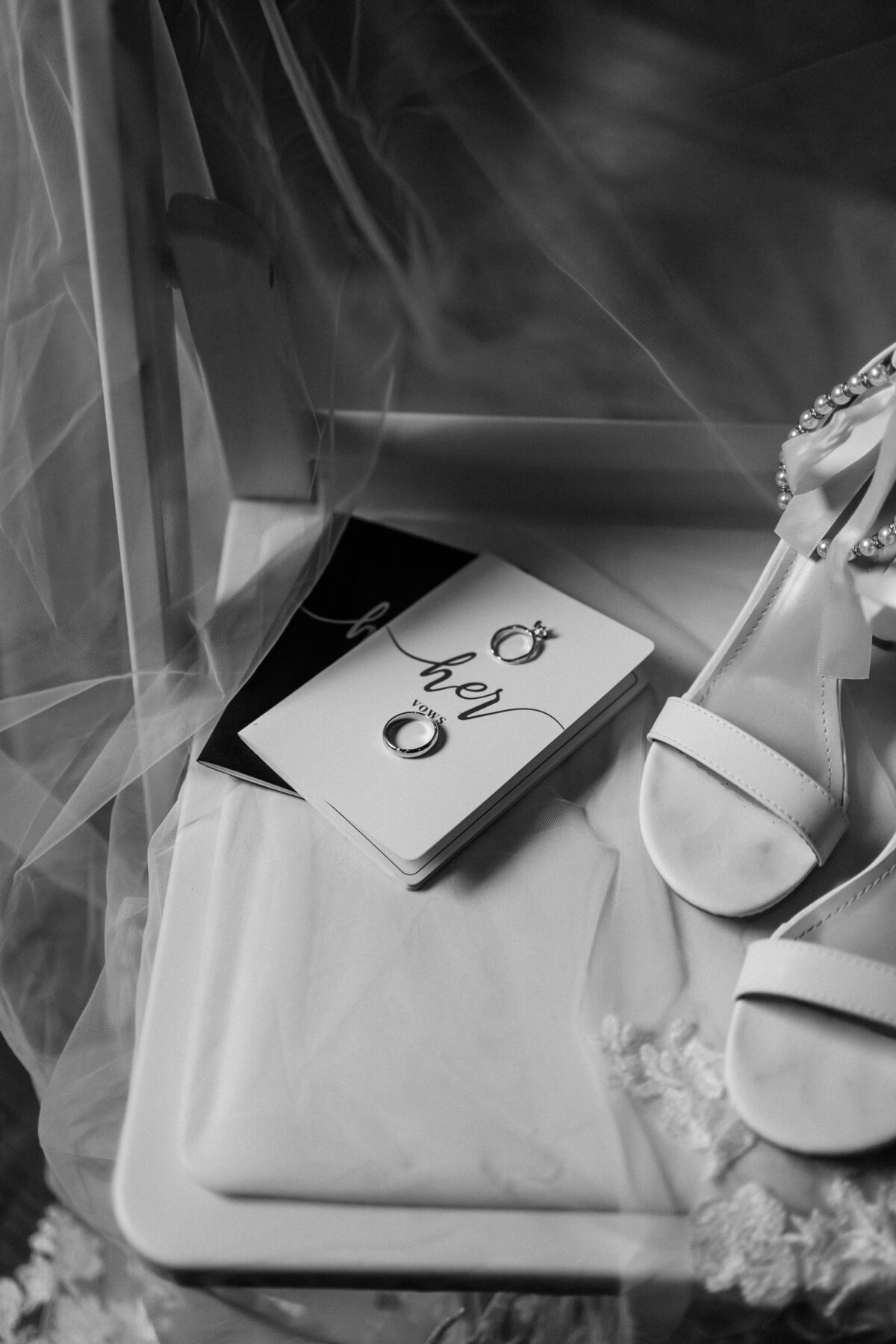 A monochrome flatlay of wedding details, including bridal shoes, "Her" vow books, and wedding rings, elegantly arranged at The Gentry Farm in Danville, Virginia, photographed by Quinn Photography LLC.