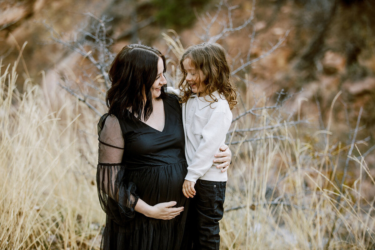 a pregnant mom in a black dress snuggles with her son while standing in front of tall, dry grass at a park in lyons, colorado