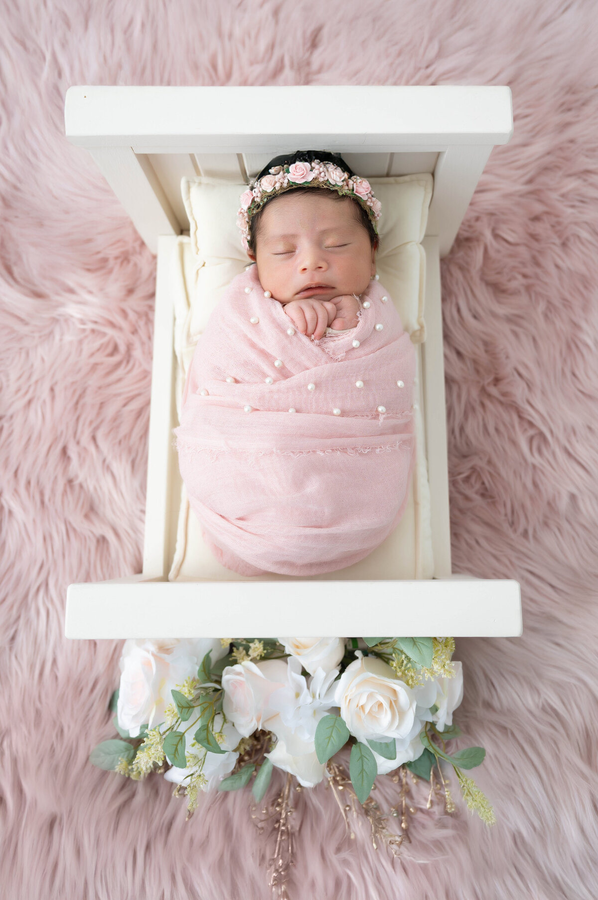 Wrapped newborn baby lying on an off-white bed placed on a pink shaggy rug, with off-white roses near the footboard.