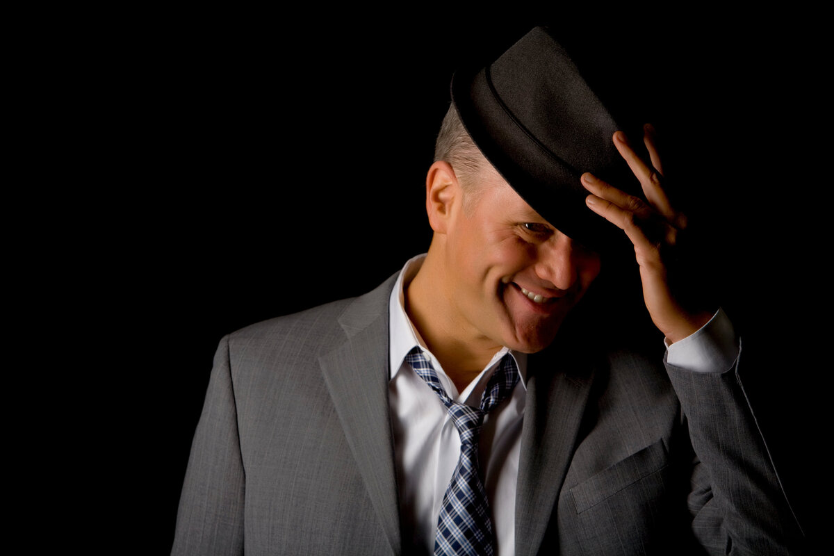 A male dating portrait client in the studio tipping his hat, smiling & wearing a grey suit