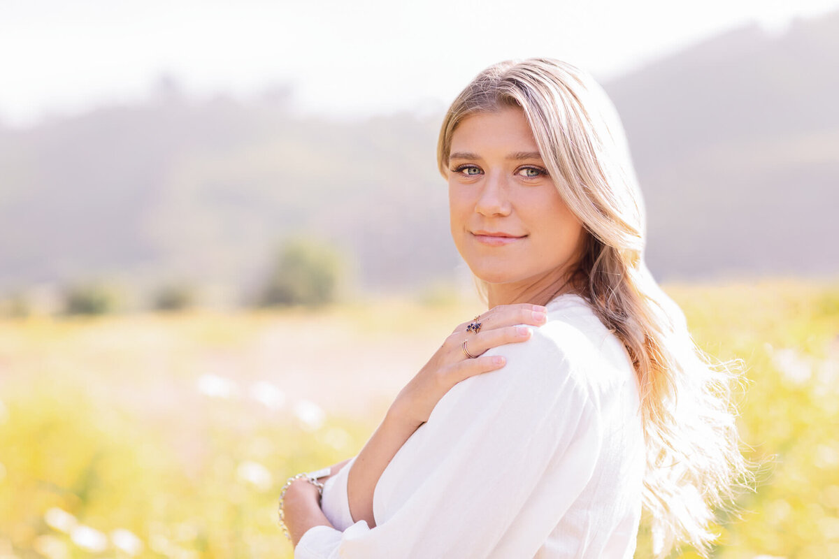A woman with long blonde hair stands in a sunlit field, wearing a white top. She has a gentle expression and holds her hands near her shoulder. The background shows blurry, green hills and a bright, clear sky.