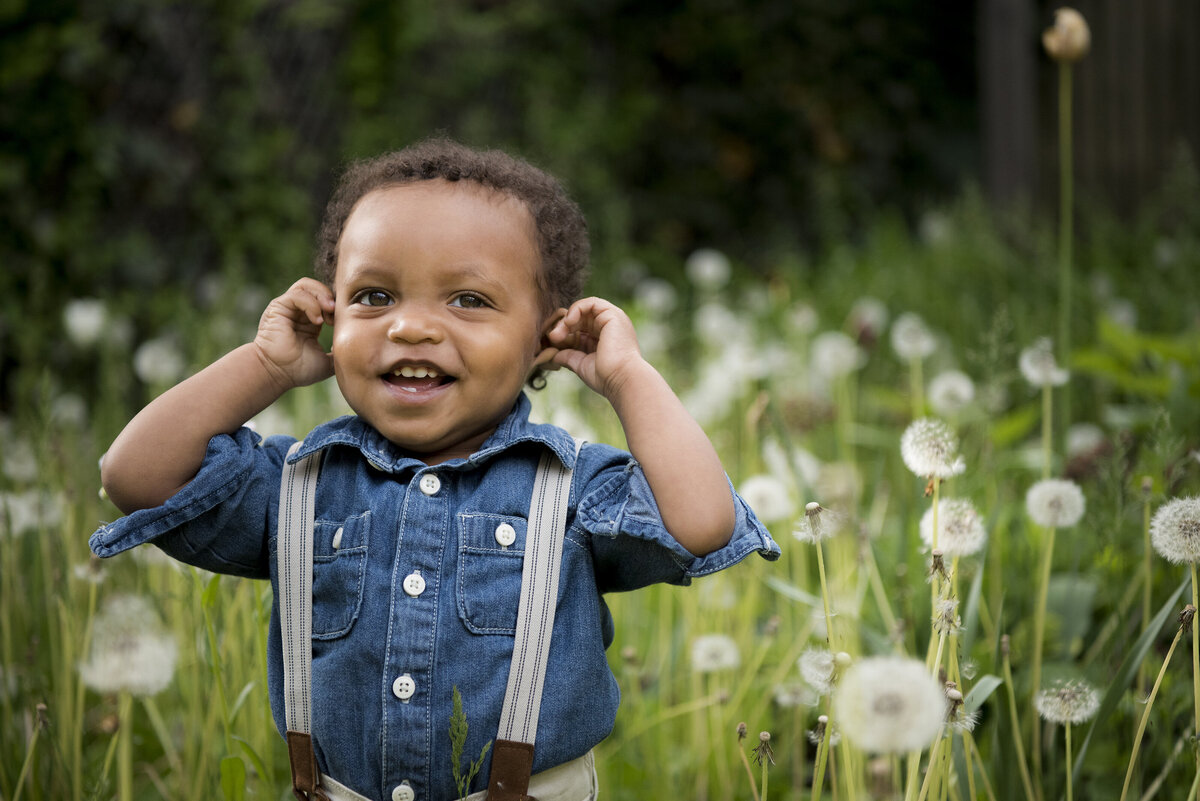 A small child smiling in a field and holding their ears 