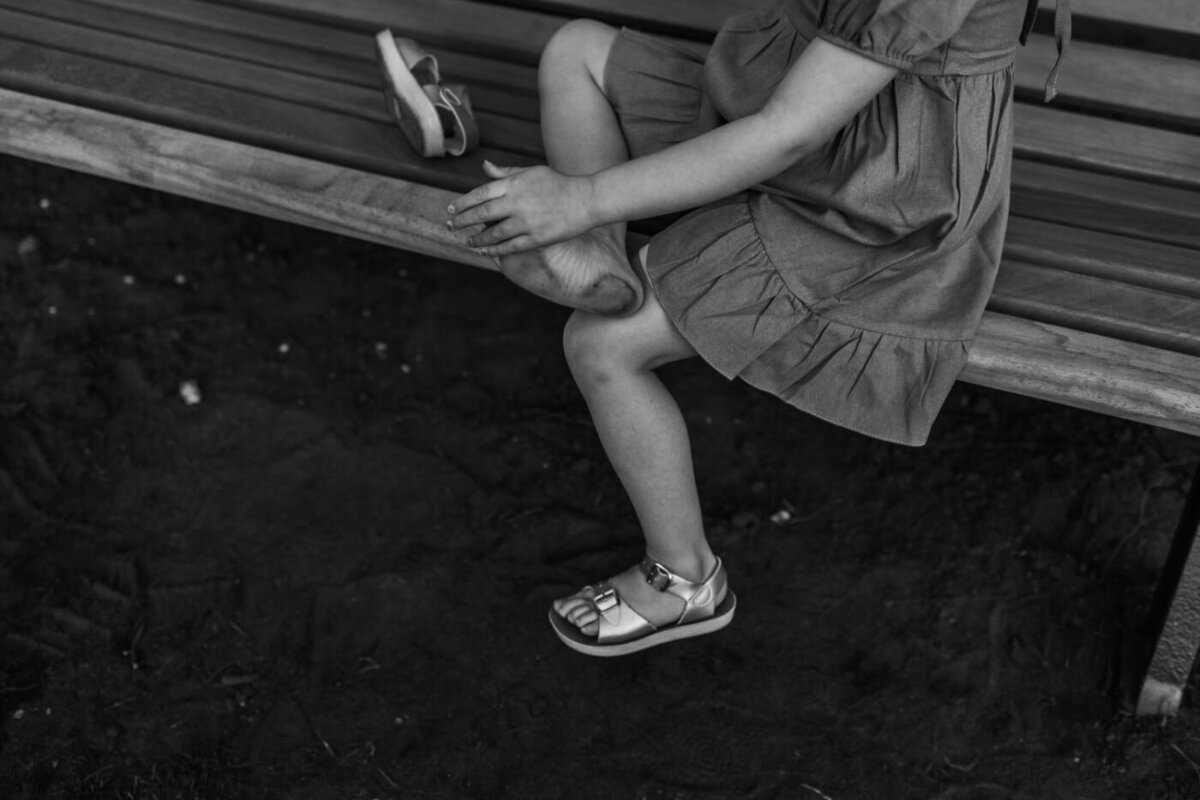 eaturing a child on a bench, focused on putting on a shiny Salt Water Sandal Surfer. The other sandal lies beside them.