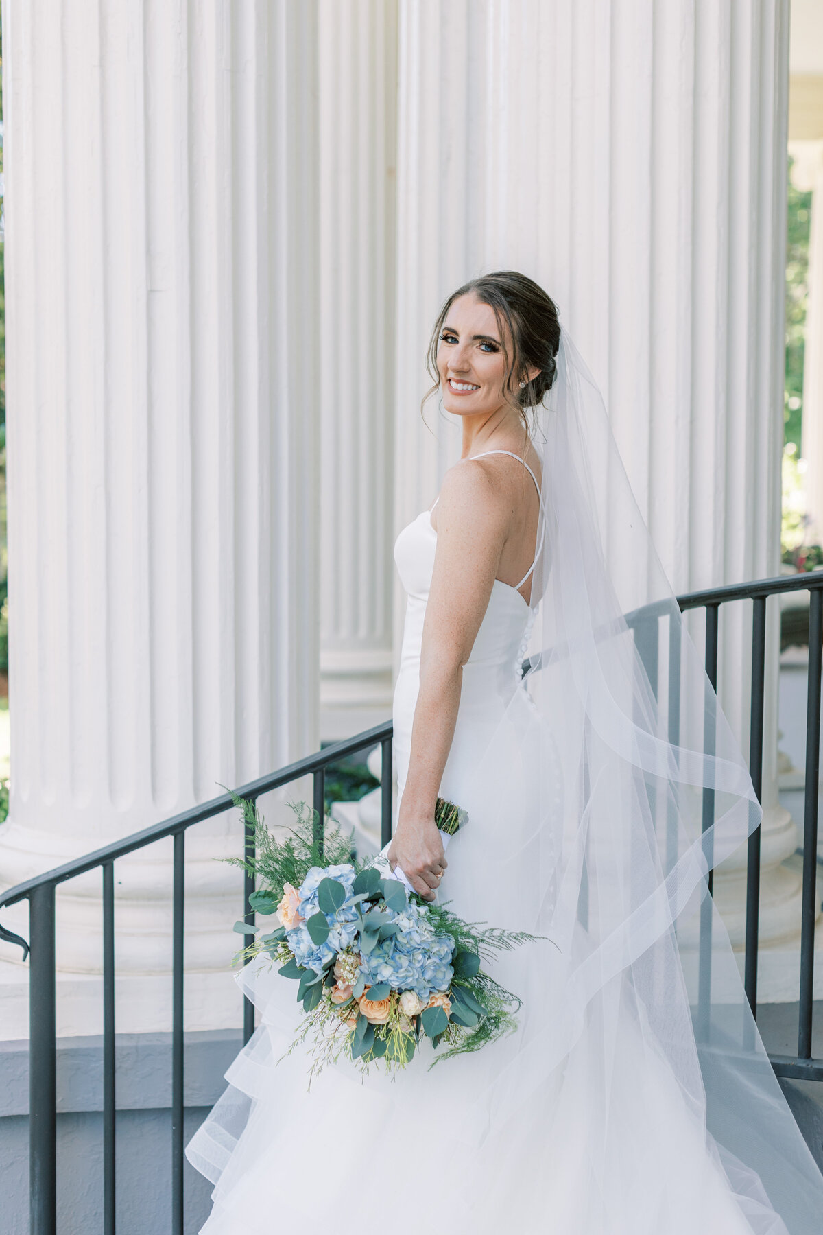A bride smiles over her shoulder while standing by a white column.