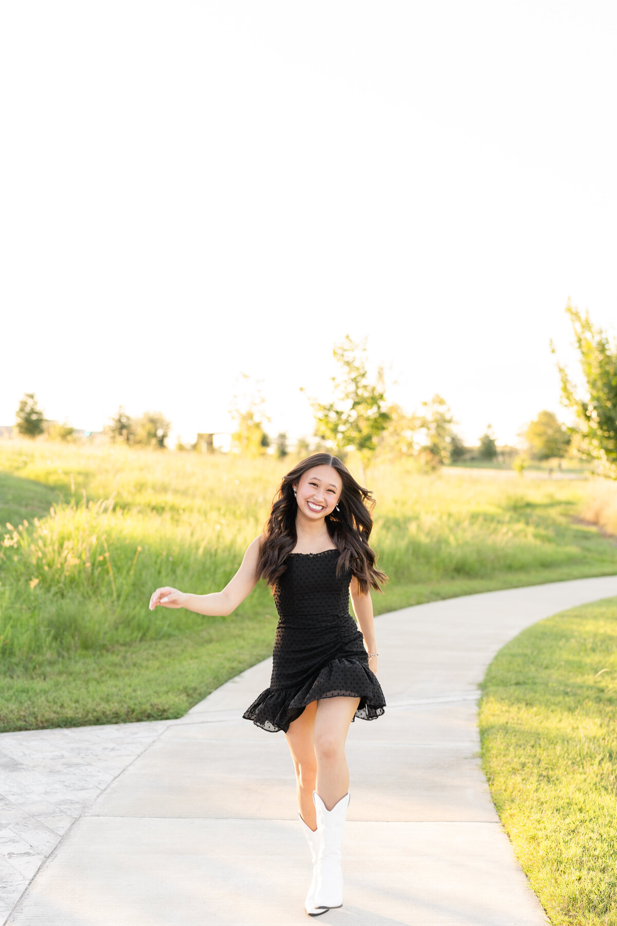 Houston High School senior girl laughing and skipping down path while wearing black dress and white boots at Josey Lake Park