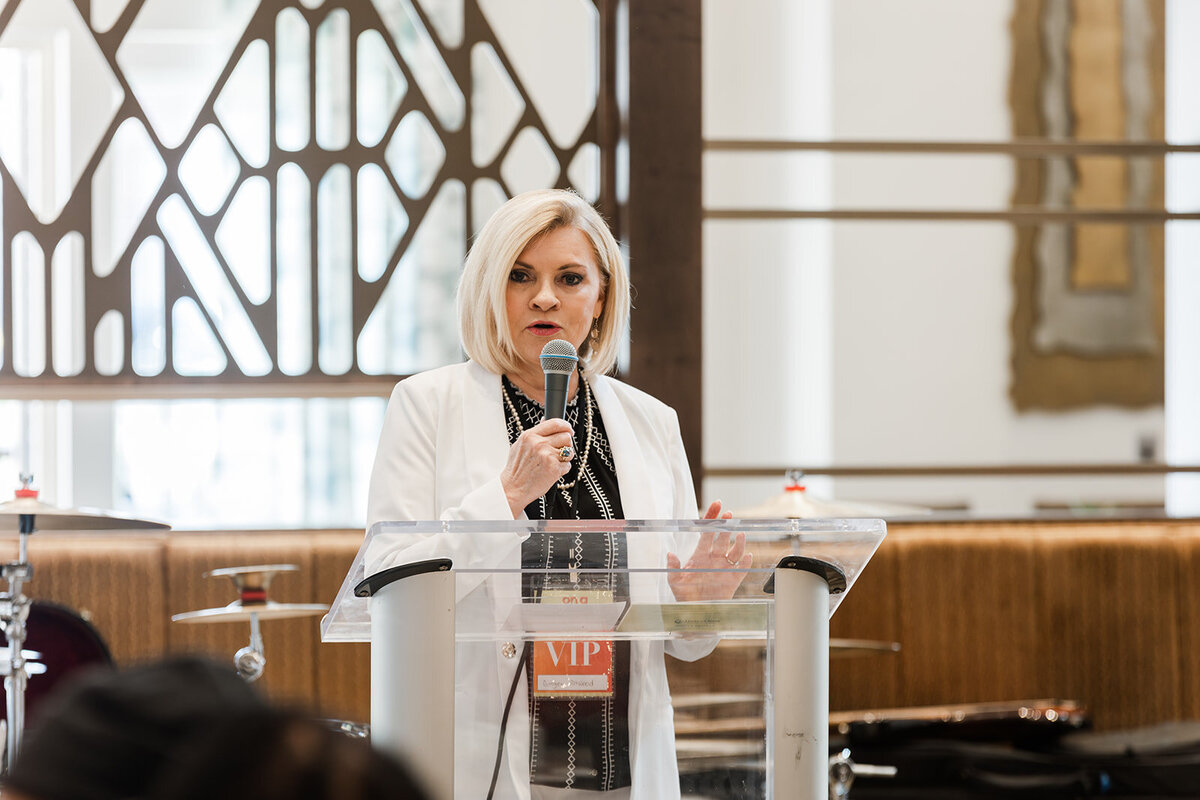 A woman speaking into a microphone at a podium in a room with modern decor and attendees in the foreground.