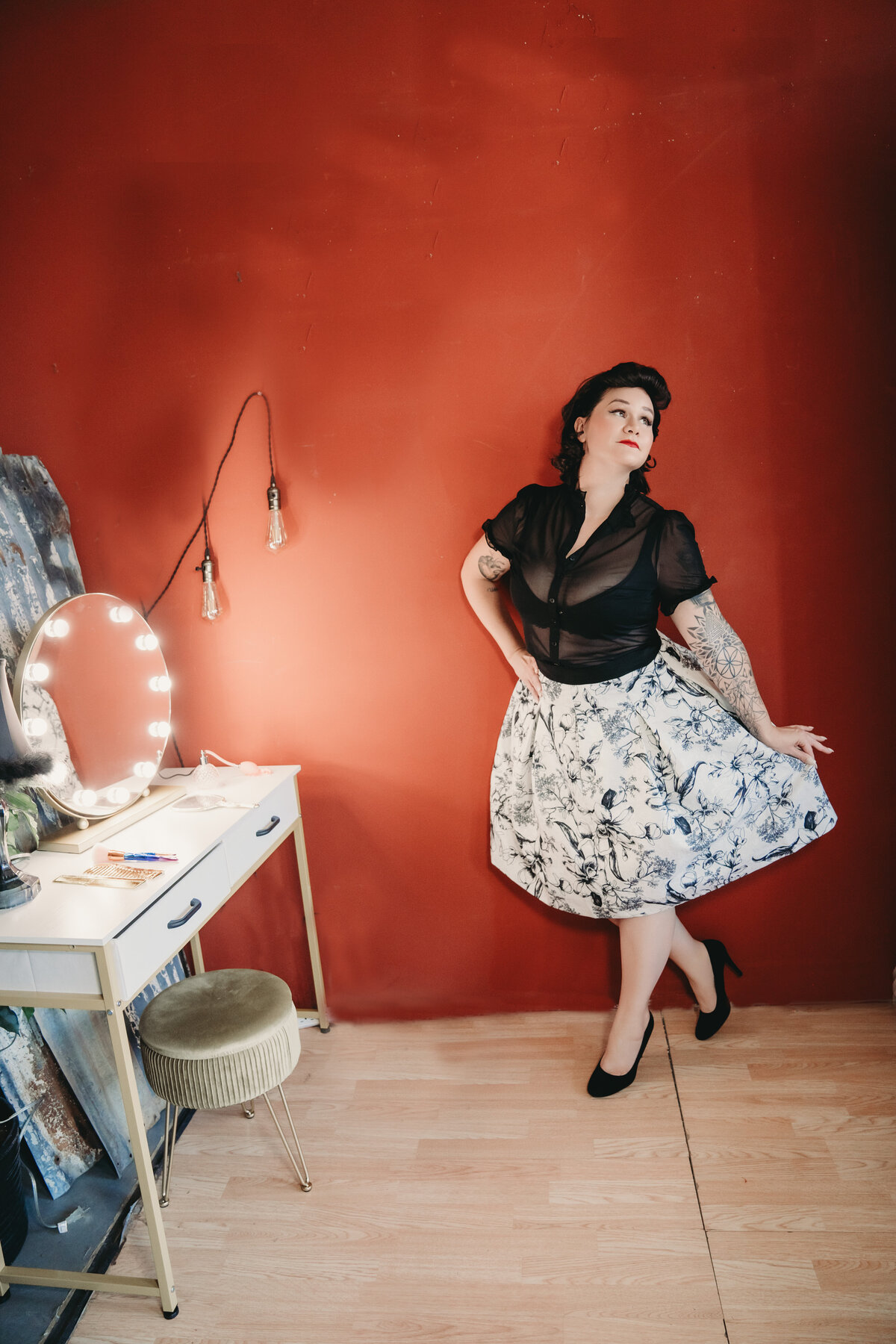 red wall with pinup woman standing in front  of it in black and white skirt and black top