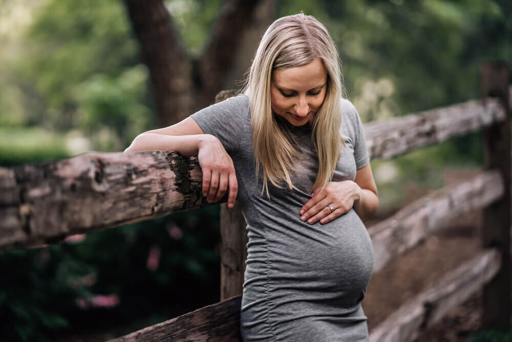 A pregnant mom stands in front of a rustic fence at Lake Harriet Lyndale Park in Minneapolis, as she gazes down at her growing baby bump.