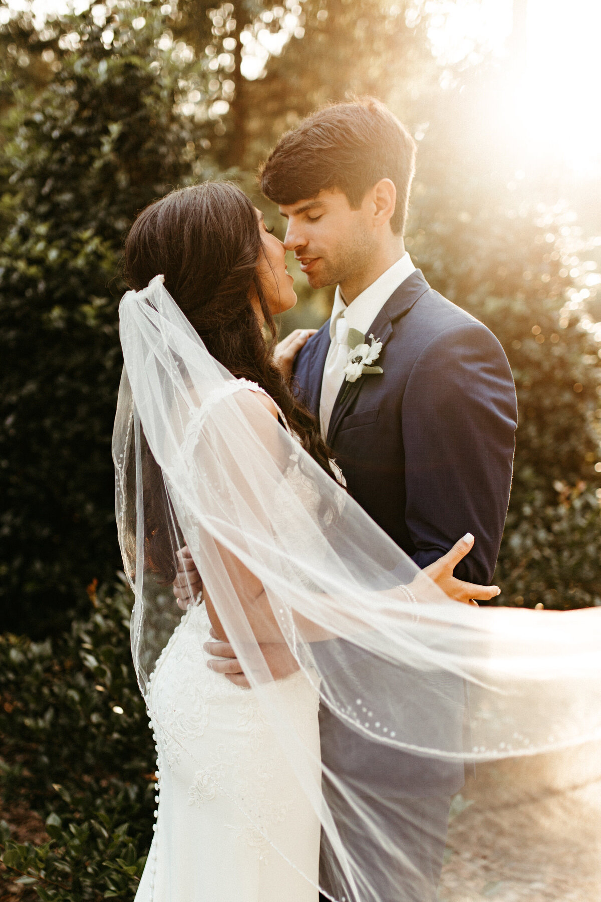 Groom holding bride close as her veil blows in the wind with a sun flare behind them