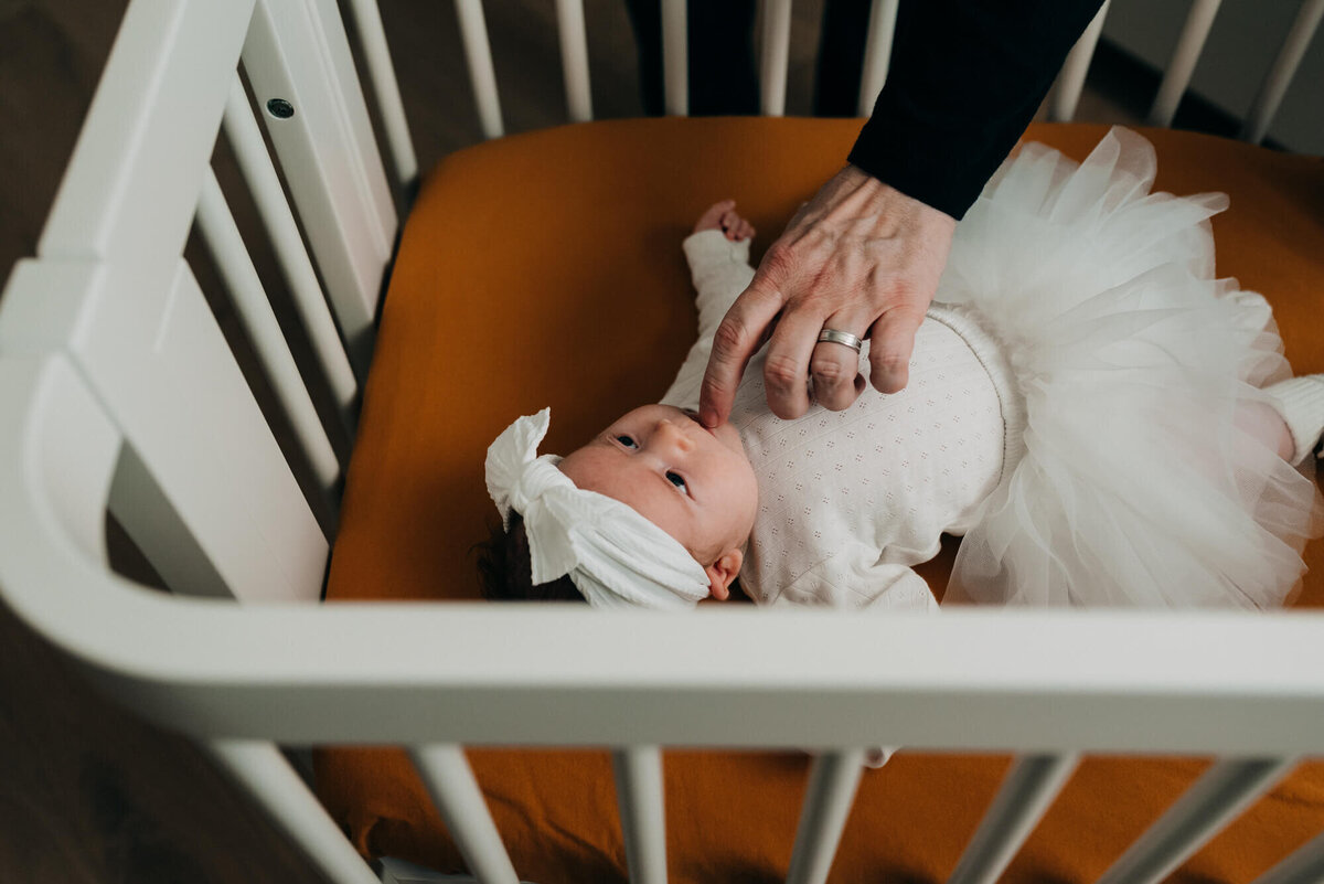 A father gently taps his daughter on her lips as she lays in her crib while wearing a white onesie and white tutu