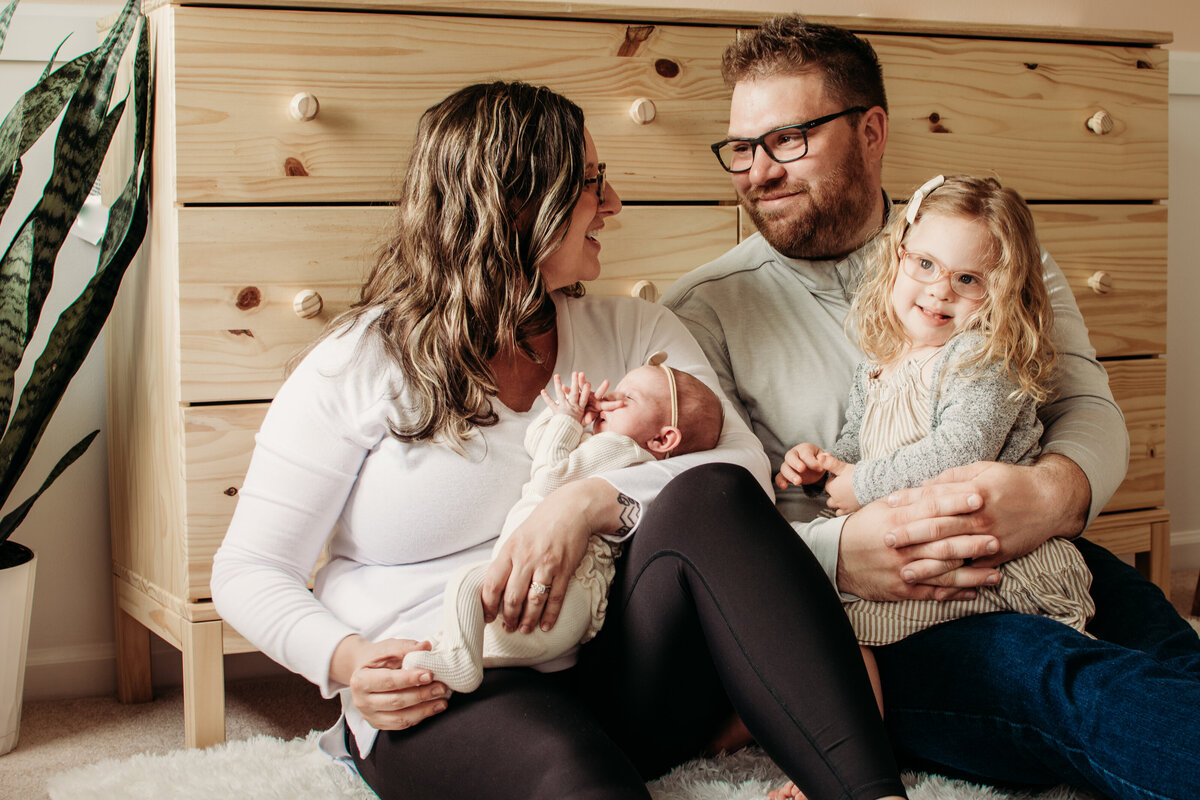 Family cuddling in the nursery by the dresser with their toddler and newborn girls during a newborn photoshoot at home in Bartlett, IL