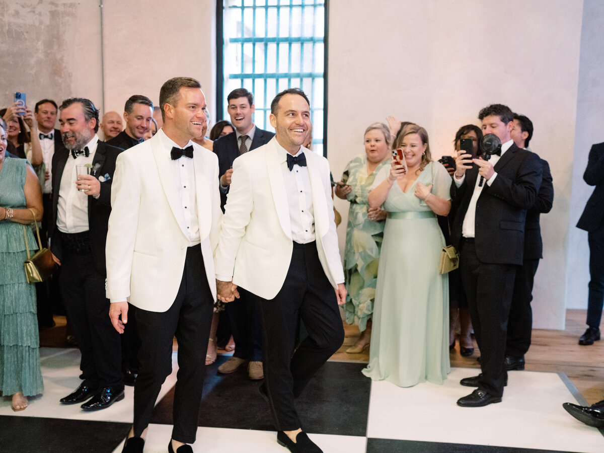 The couple joyfully walks hand in hand into their wedding celebration, greeted by friends and family, embracing the love and support surrounding them on their special day.