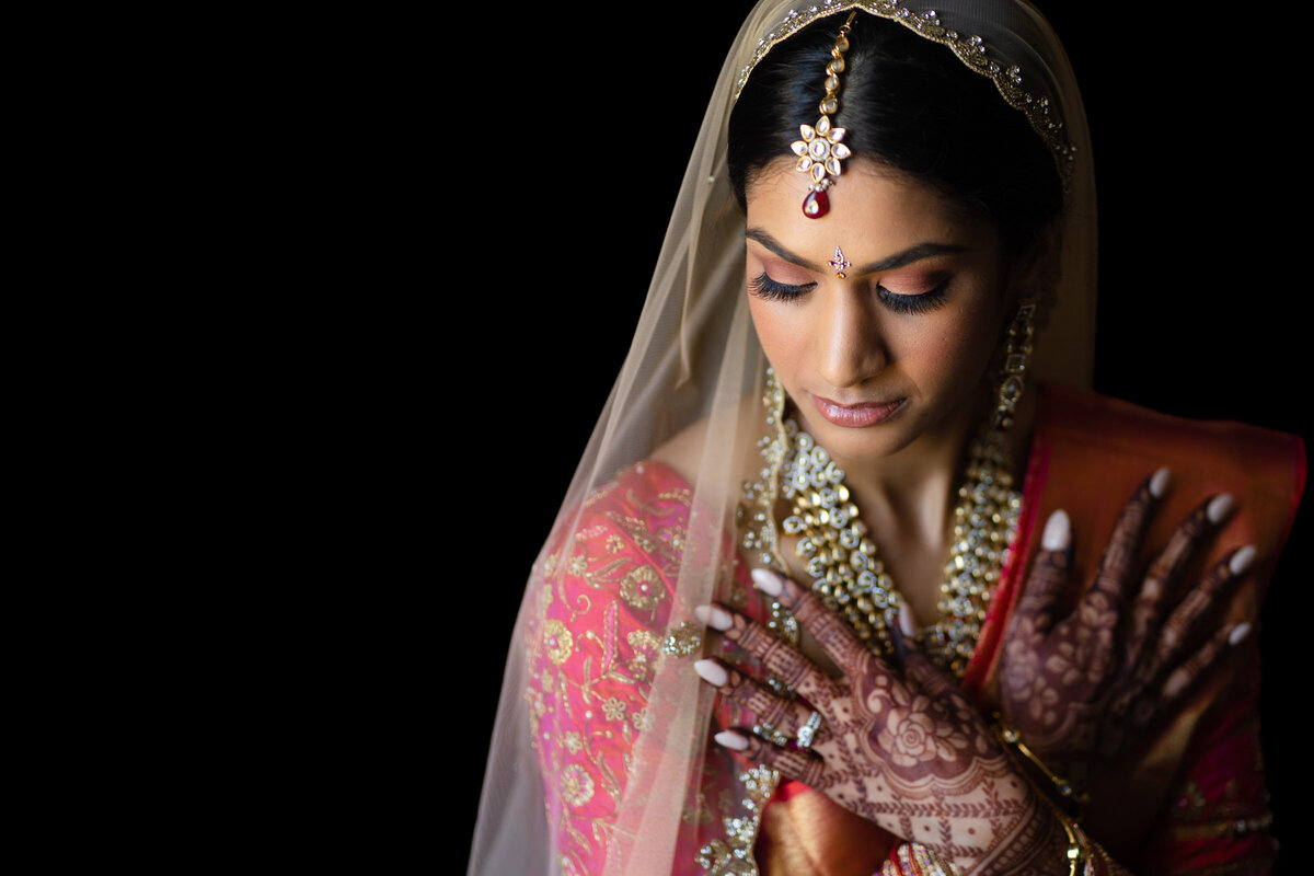 A South Asian bride in traditional attire closes her eyes, her hands decorated with intricate henna designs