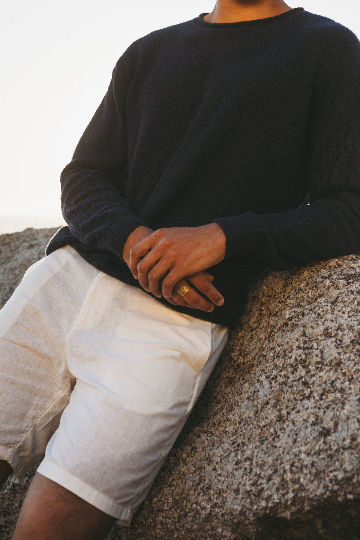 Close-up of Timothe’s hands resting on a rock, wearing a navy sweater and white linen shorts during golden hour in Cape Town.