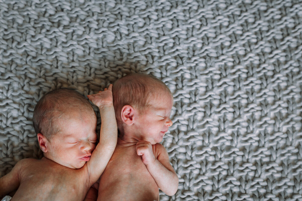twin newborns with no shirts sleeping with arms draped on each other on textured blanket