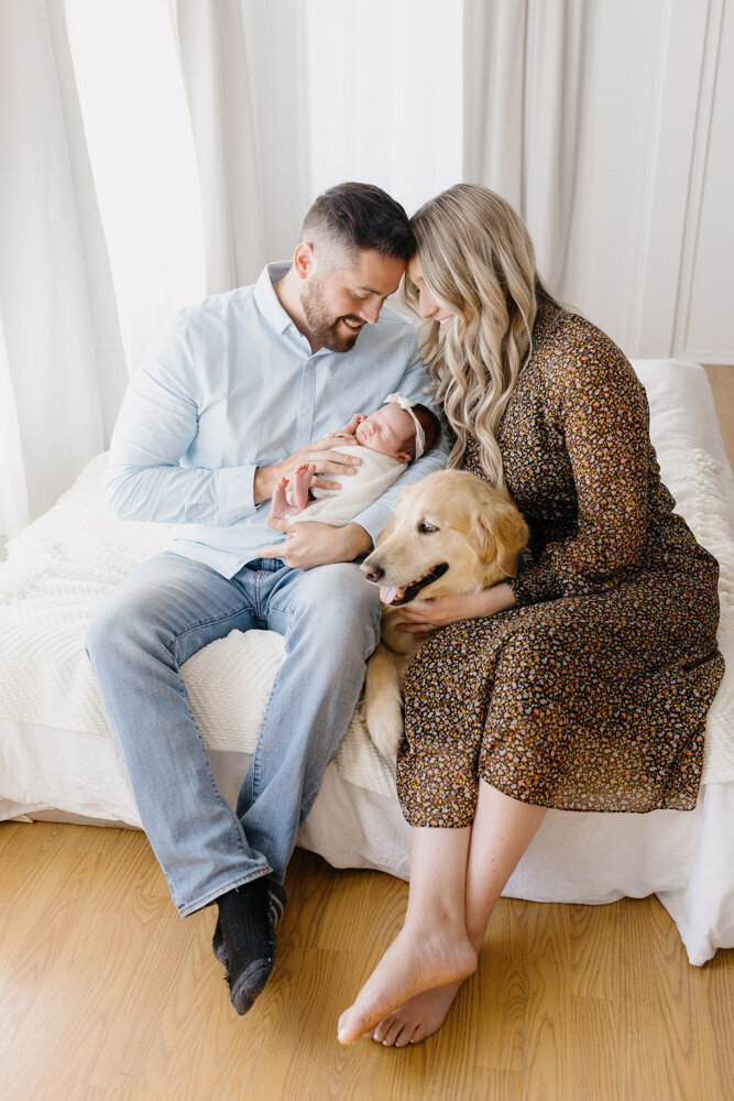parents and newborn baby sitting on bed cuddled in close admiring her with their golden retriever in Los Osos newborn photography studio