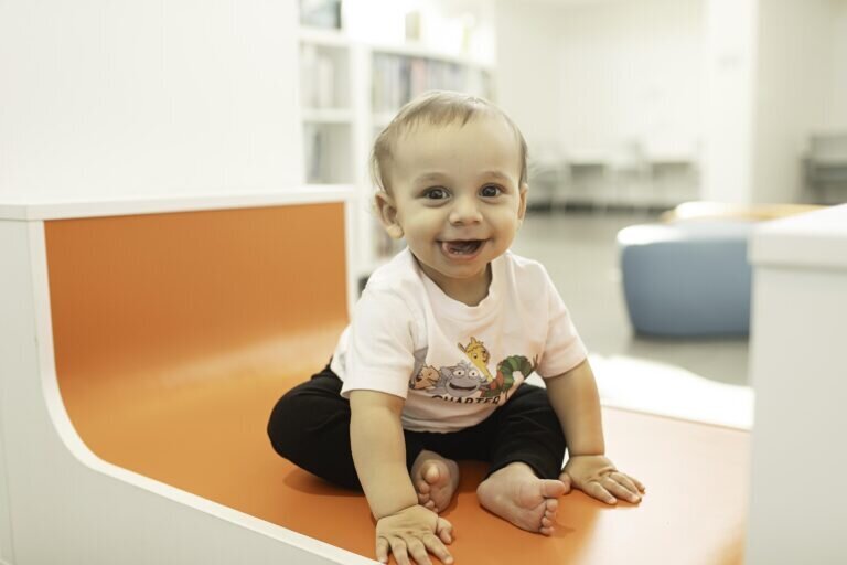 One-year-old child sitting on the floor of a library, surrounded by colorful books, looking curious and happy