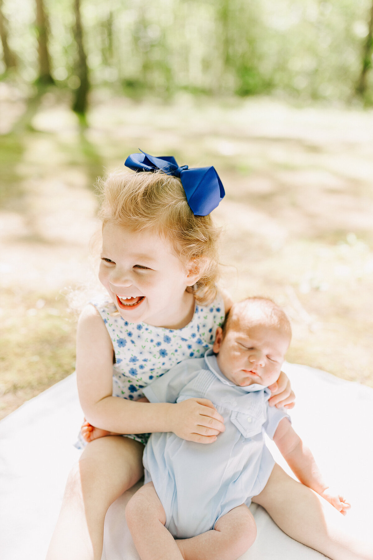 A proud big sister with curly red hair and a bright blue bow grins while holding her newborn baby sibling in a soft, natural outdoor setting. This sweet sibling moment captures the joy and tenderness of welcoming a new baby, reflecting the light and heartfelt style of a Birmingham newborn photographer.