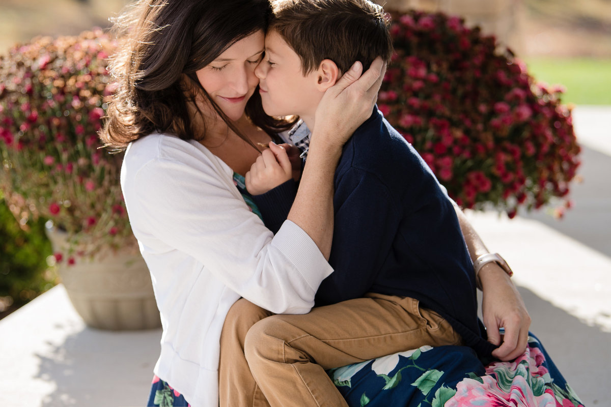 little boy sitting on mother's lap snuggling in front of potted mum plants