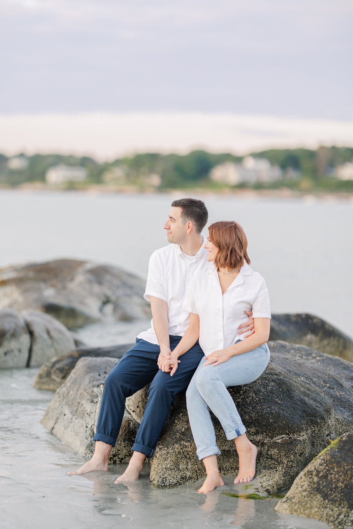 A couple holding hands and watching the sunset as they sit on a rock during their Wingaersheek Beach engagement pictures.