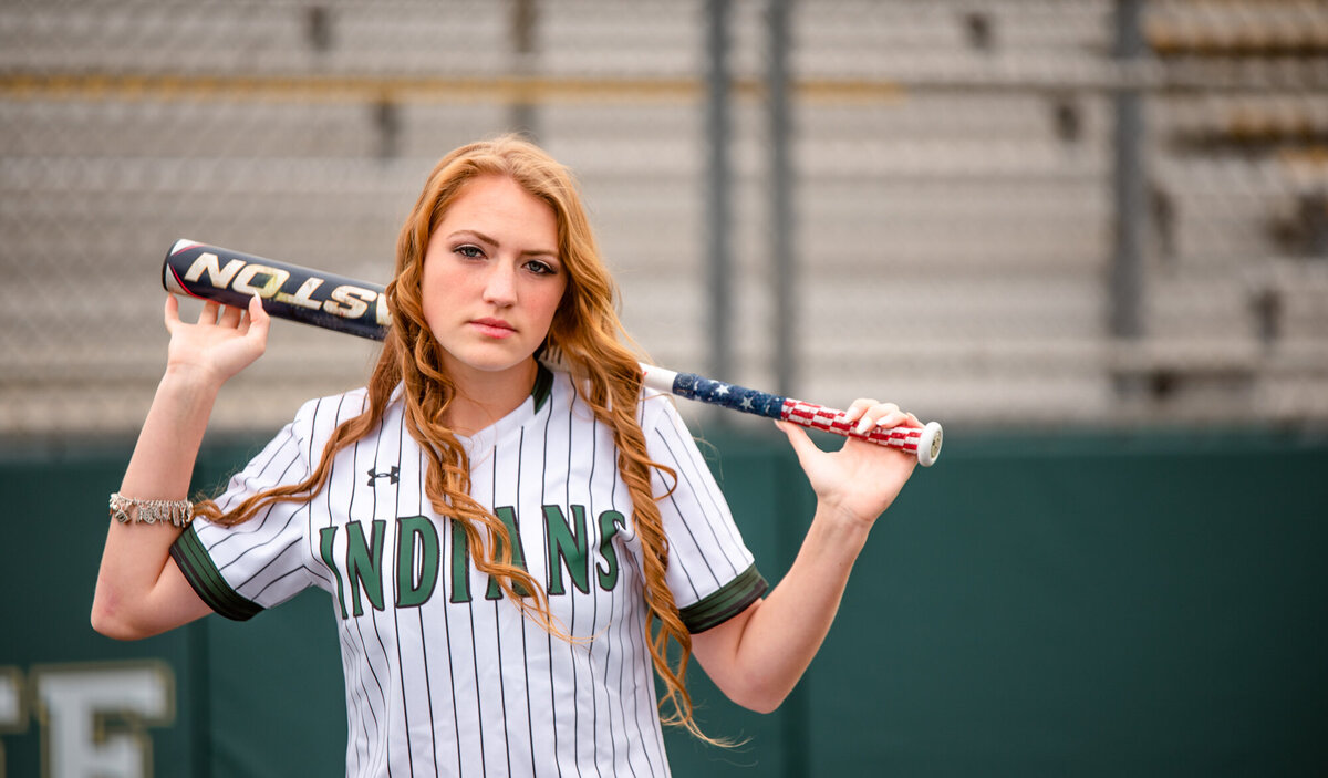 A recent graduate stands near home plate holding her softball bat behind her head while looking at the camera.