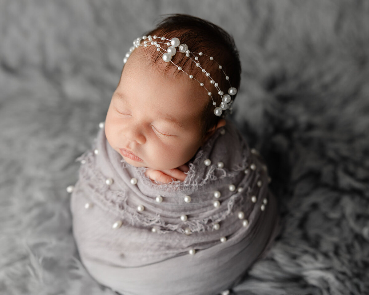 Newborn baby girl delicately wrapped in gray fabric adorned with pearls, with a matching pearl headband, resting peacefully on a soft gray backdrop. Captured by Inland Empire newborn photographer Kaitlyn Dawn Photography.