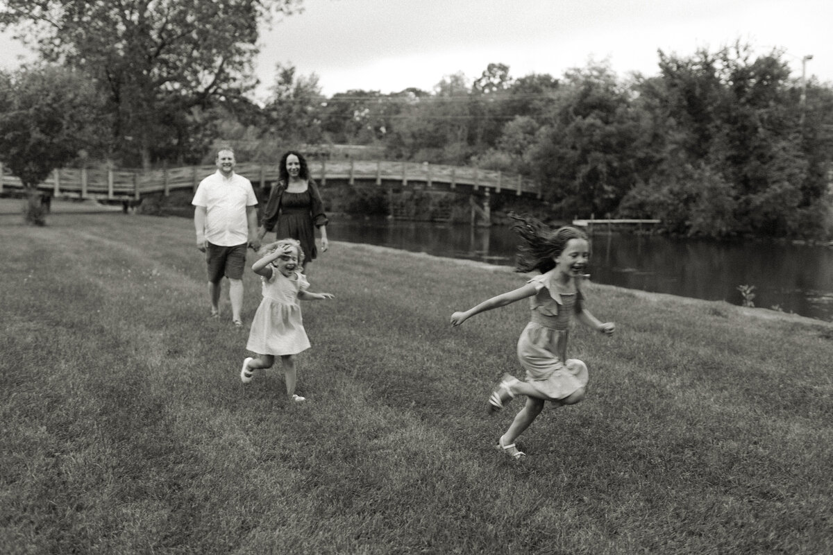 A mom and dad stroll hand-in-hand while their daughters play tag during playful family pictures captured by Emmie Verdell Photo.