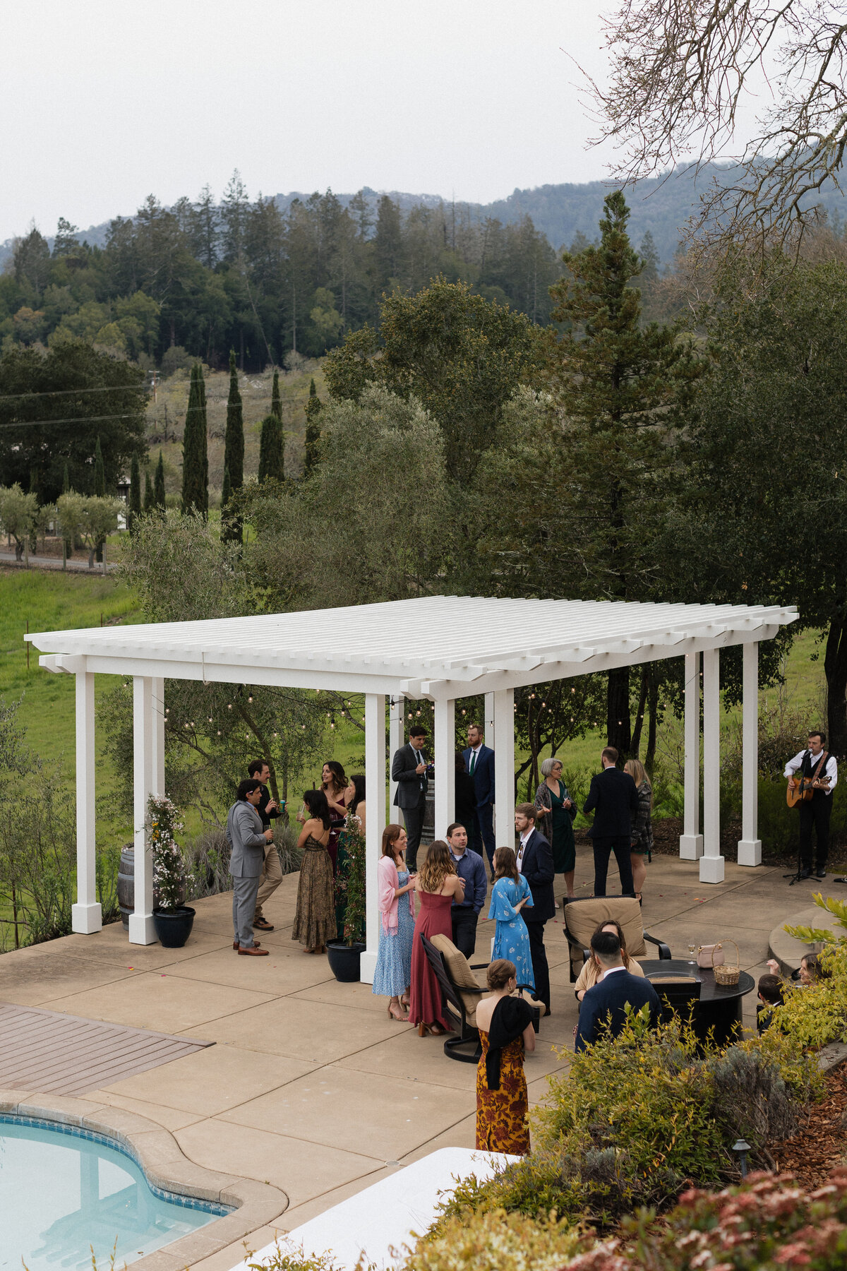 Guests enjoying an outdoor wedding cocktail hour under a white pergola, surrounded by lush greenery and scenic rolling hills.