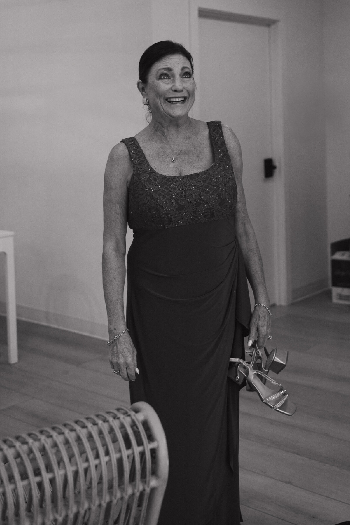 A older women looks up emotionally holding chunky low heels in one hand and a tissue in her other hand. She is in a floor length mother of the bride dress with a lace and beaded top and a flowey bottom. 