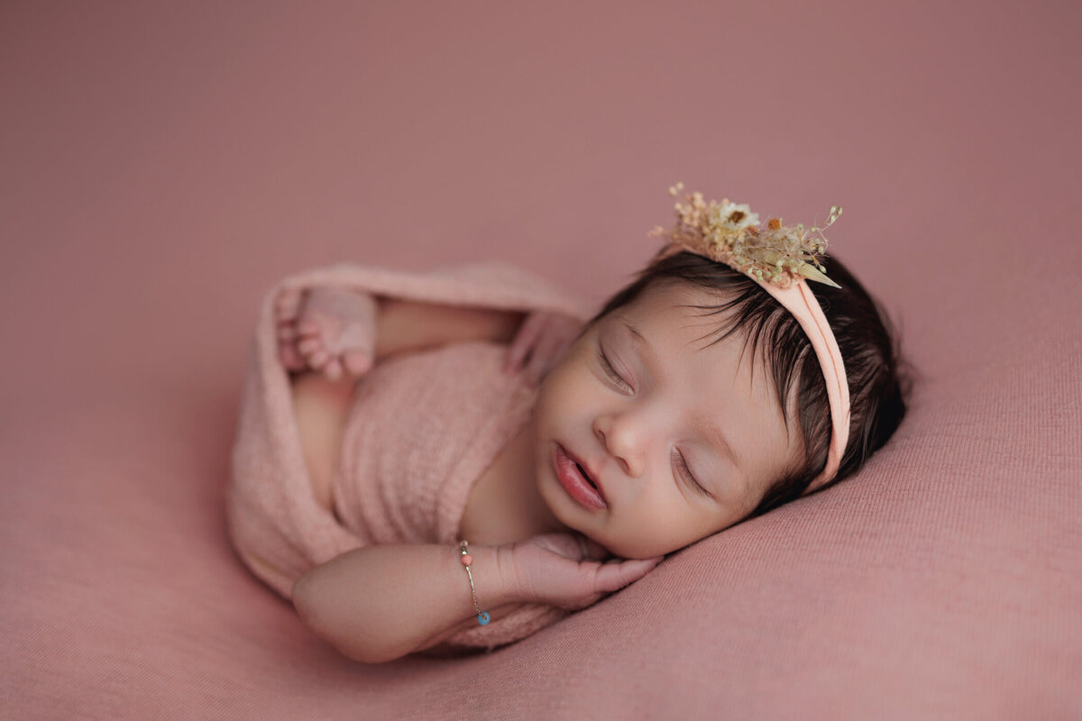 Newborn baby girl peacefully sleeping on a soft pink blanket, wrapped in a pink swaddle. She wears a delicate floral headband and a tiny bracelet on her wrist.