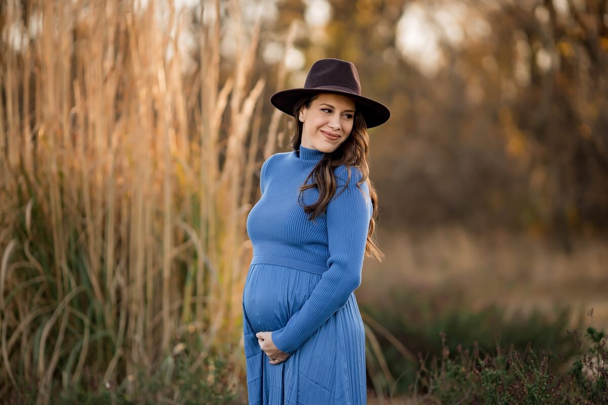 A young pregnant woman in a blue dress and hat poses for maternity photos in a field.