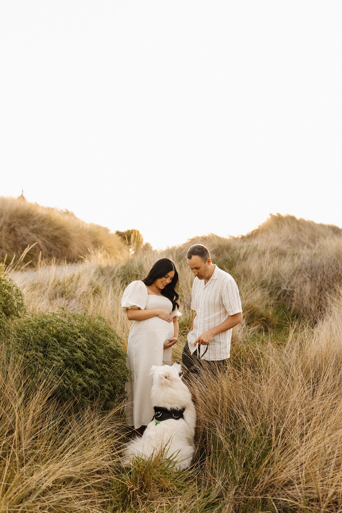 A beautiful mother to be and her husband and their dog standing in the long grass at sunset and looking at one another while lucy jane photography takes candid photographs of them in christchurch