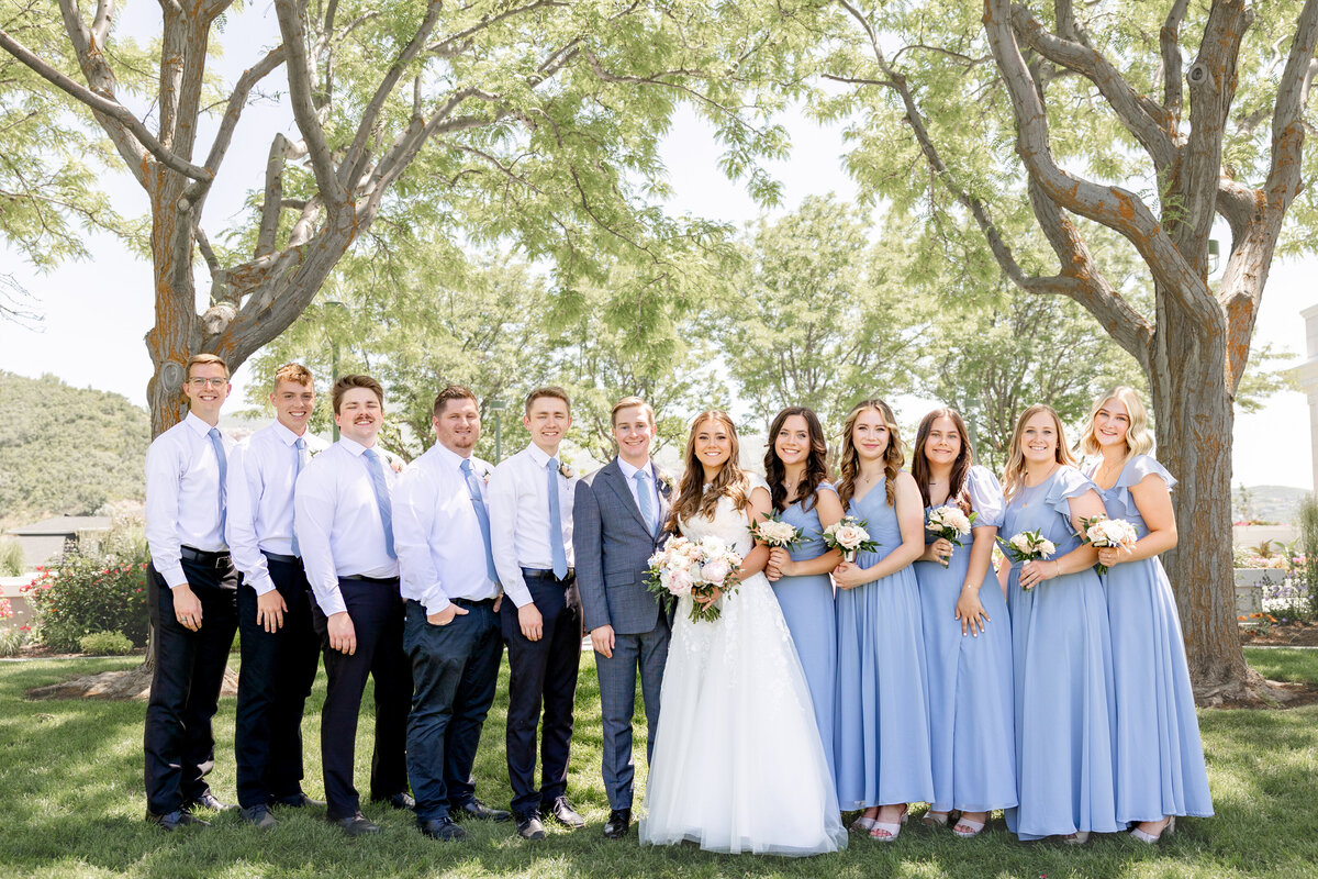 The bride, groom, bridesmaids, and groomsmen pose under lush green trees near the Bountiful Utah LDS Temple.