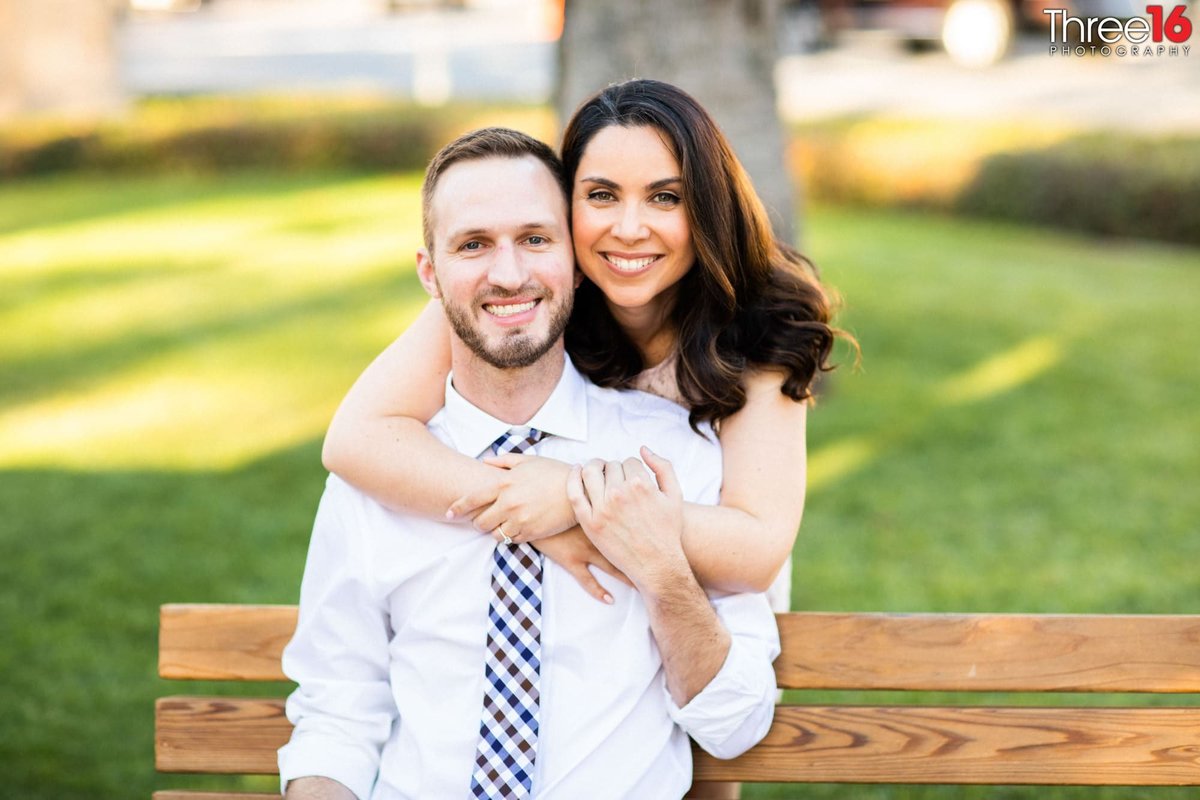 Bride to be embraces her Groom from behind as he sits on a park bench