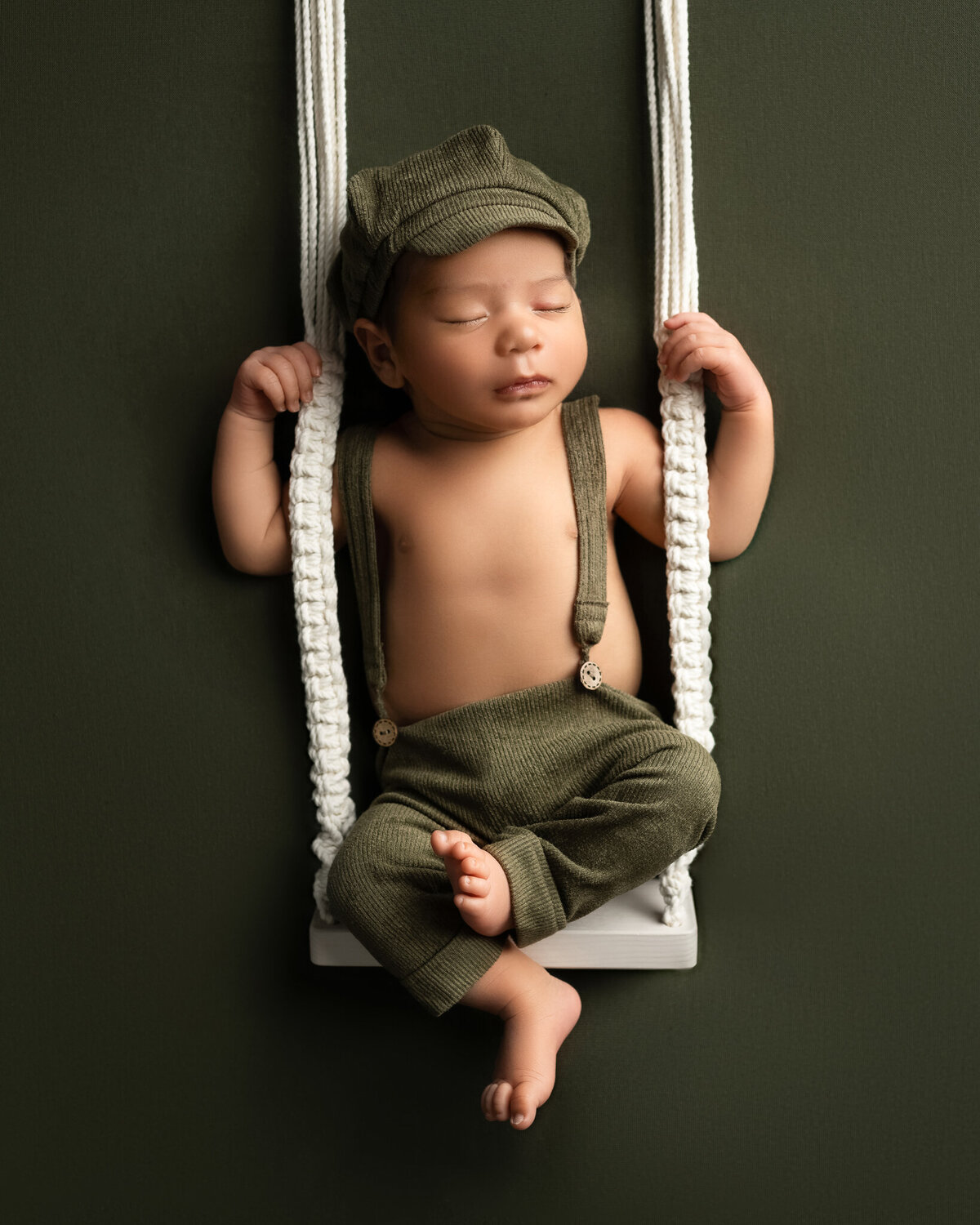Newborn boy in green corduroy outfit on suspended swing set against a dark olive backdrop | Studio newborn photography in Cedar Park and Austin