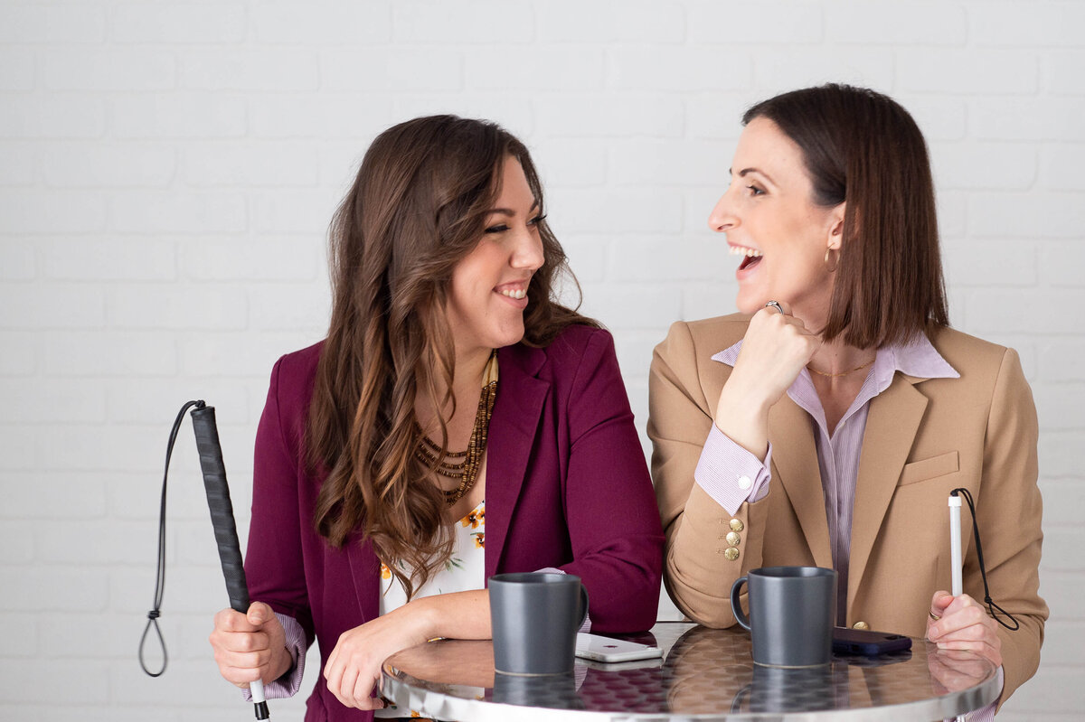 two women one in a burgundy blazer, the other in a camel blazer laughing and smiling at each other at a bar table.  Captured in studio by Ottawa Branding Photographer JEMMAN Photography COMMERCIAL