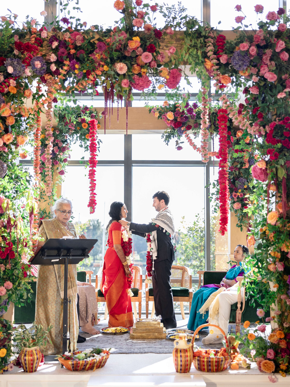 A couple stands under a vibrant floral arch during a ceremony. The woman is in a red saree, and the man wears a scarf over his suit. An officiant and two seated guests are present, surrounded by colorful flowers and traditional décor.