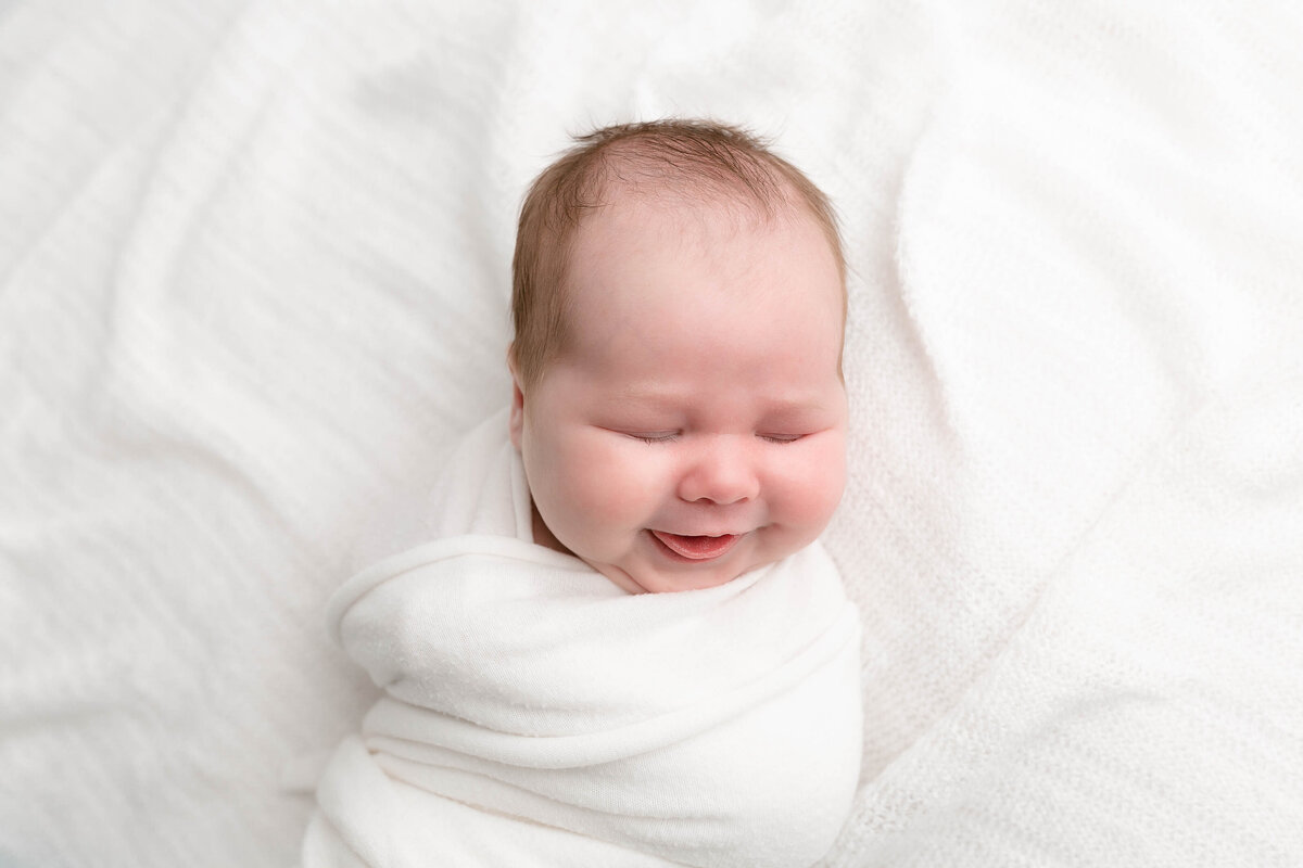 light-skinned baby wrapped in white swaddle on a white textured backdrop smiling with tongue sticking out at newborn photography session in portland oregon.