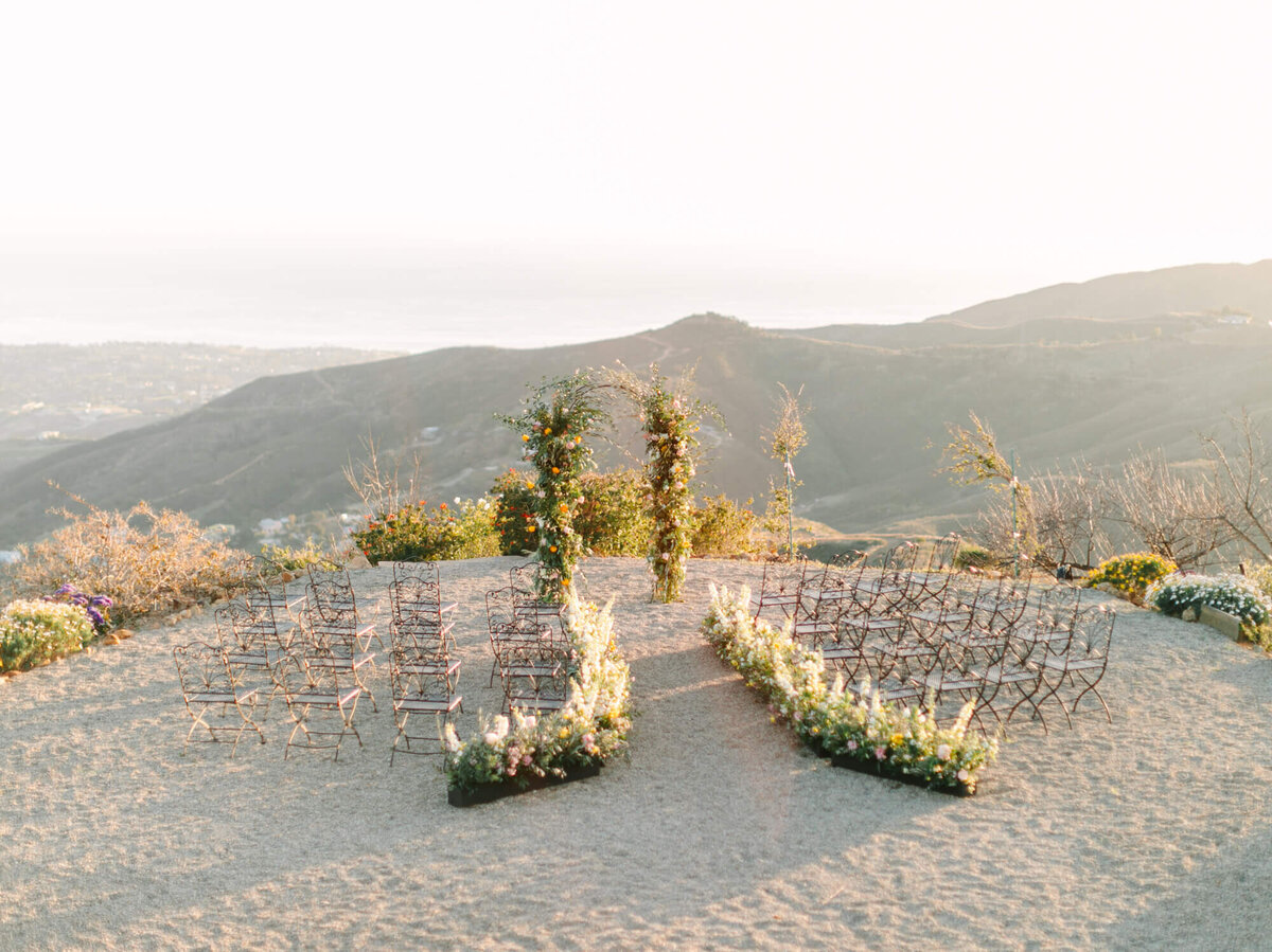 A wedding ceremony setup on a hilltop with rows of chairs and a floral arch. Soft sunlight casts a warm glow over the distant rolling hills.