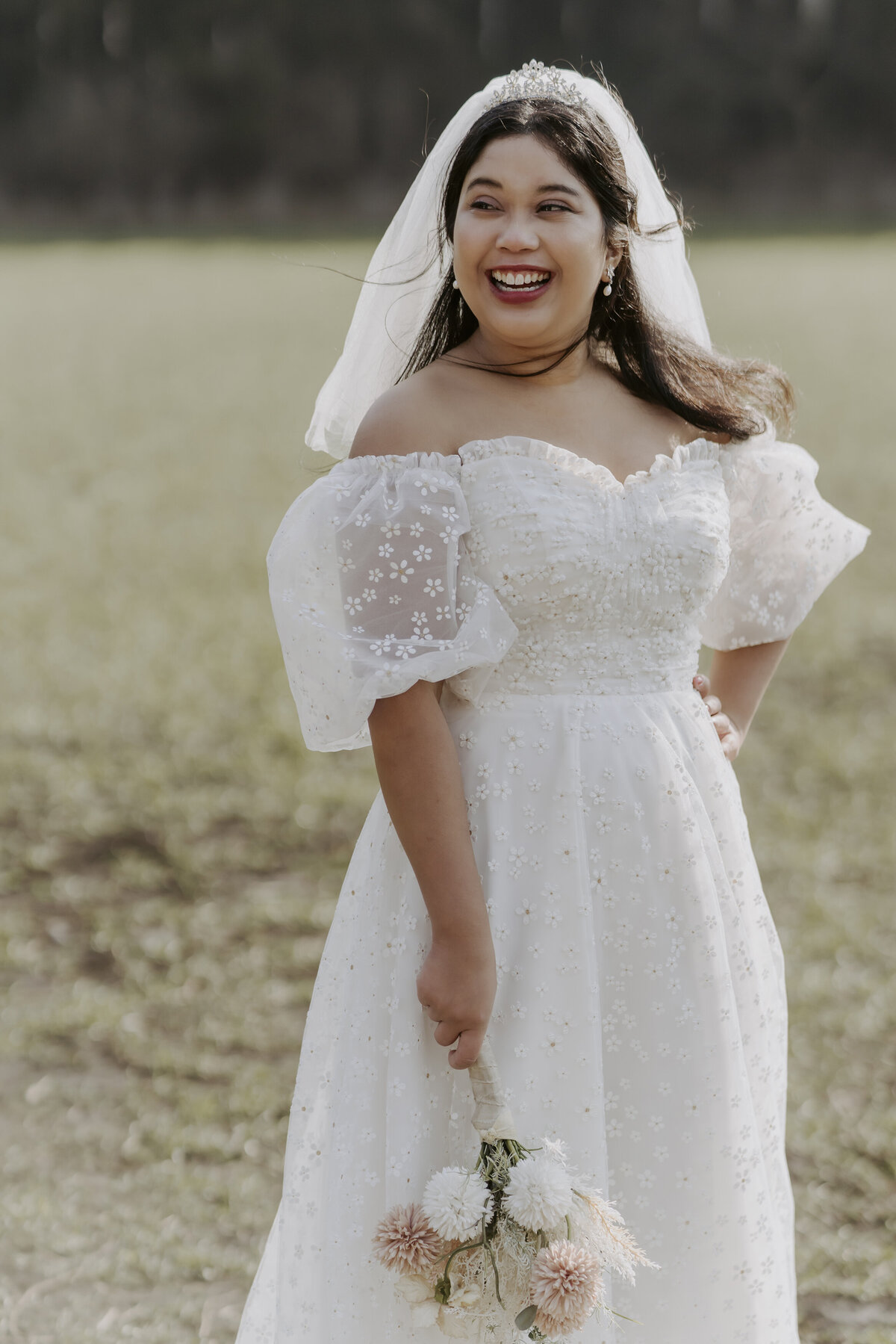 the bride is smiling and wears an off shoulder white dress and a veil while holding a bouquet