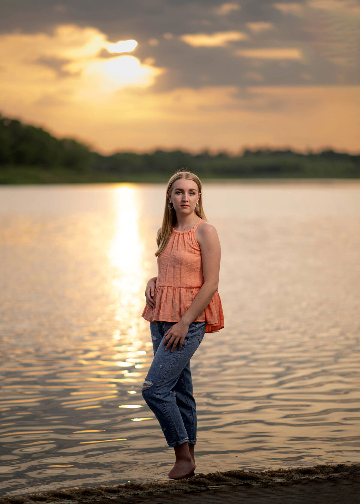 A high school senior stands in shallow lake in jeans and peach blouse at sunset