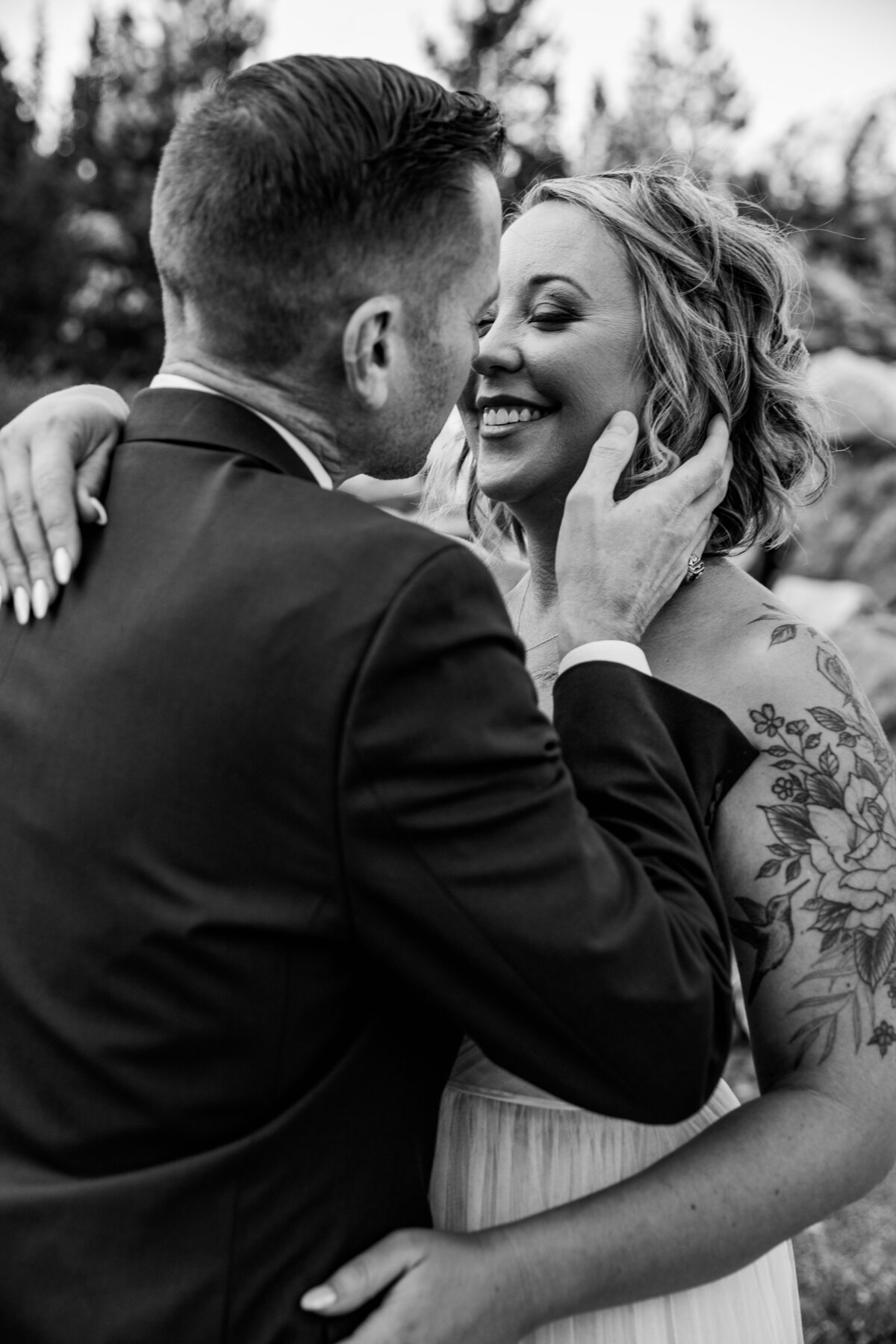 Black and white photo of a groom gently holding his bride’s face as she smiles, capturing a joyful and intimate elopement moment outdoors.