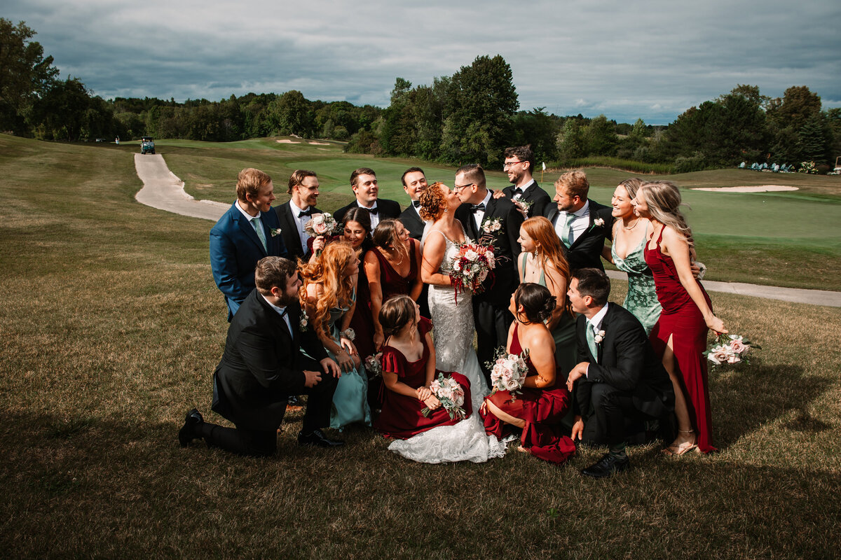 A bride and groom kiss in an open field with their large wedding party surrounding them and watching