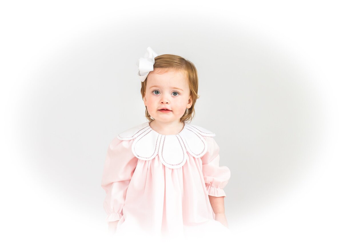 Toddler girl wearing a light pink dress with a scalloped collar and a white hair bow.