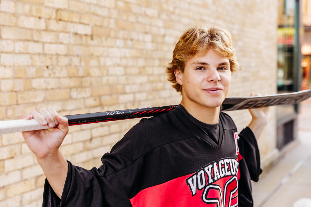 handsome young man in a hockey jersey holding his hockey stick on his shoulders as he looks over captured by Ashley Kalbus Photography.