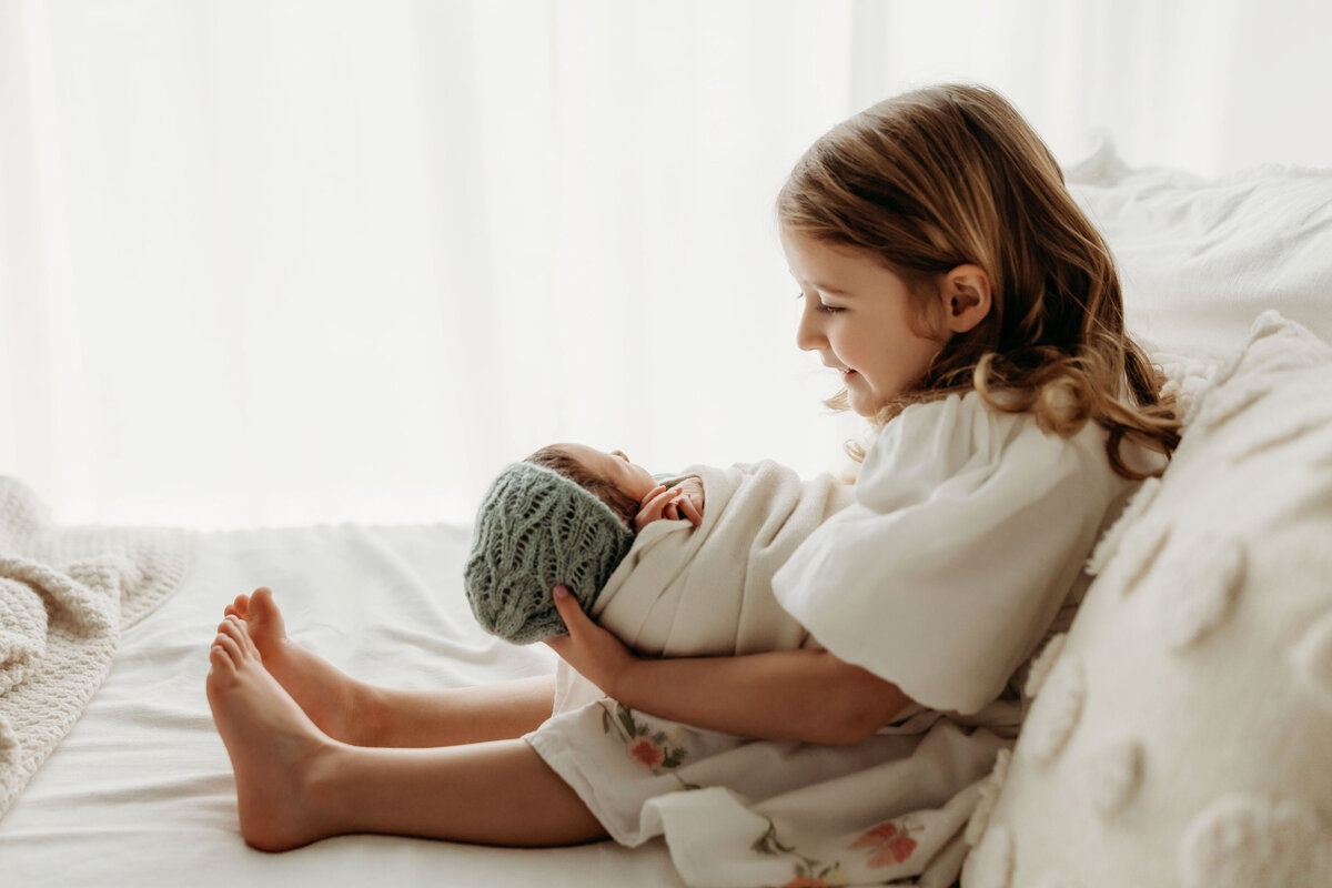 A big sister holds her newborn baby sister and admires her, inside a Vancouver Washington natural light studio